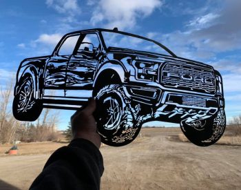 Hand holding a black metal cutout silhouette of a lifted Ford pickup truck against a rural dirt road and blue sky.