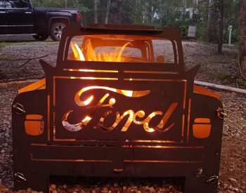 Outdoor fire pit shaped like the front of a vintage Ford truck with flames glowing through the grille at a wooded campsite.