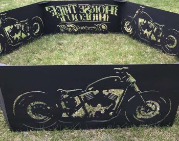 Large motorcycle-themed metal fire pit ring with cutout biker graphics and text, displayed on grass.
