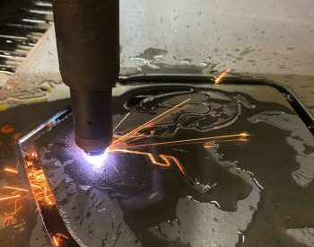 Plasma torch cutting a detailed design into a steel plate, with bright sparks and molten metal.