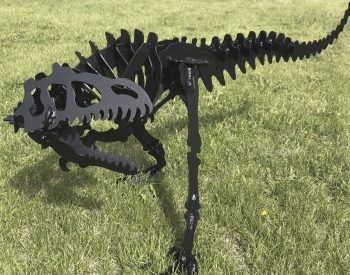 Black metal dinosaur skeleton sculpture standing on grass, made from layered steel plates.